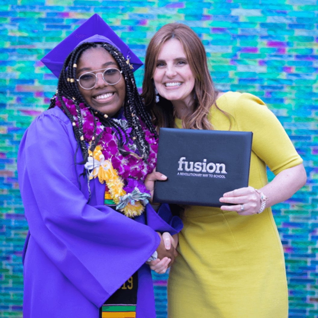 Student in cap and gown on the left shakes hand with teacher on the right while both hold a Fusion Academy diploma.